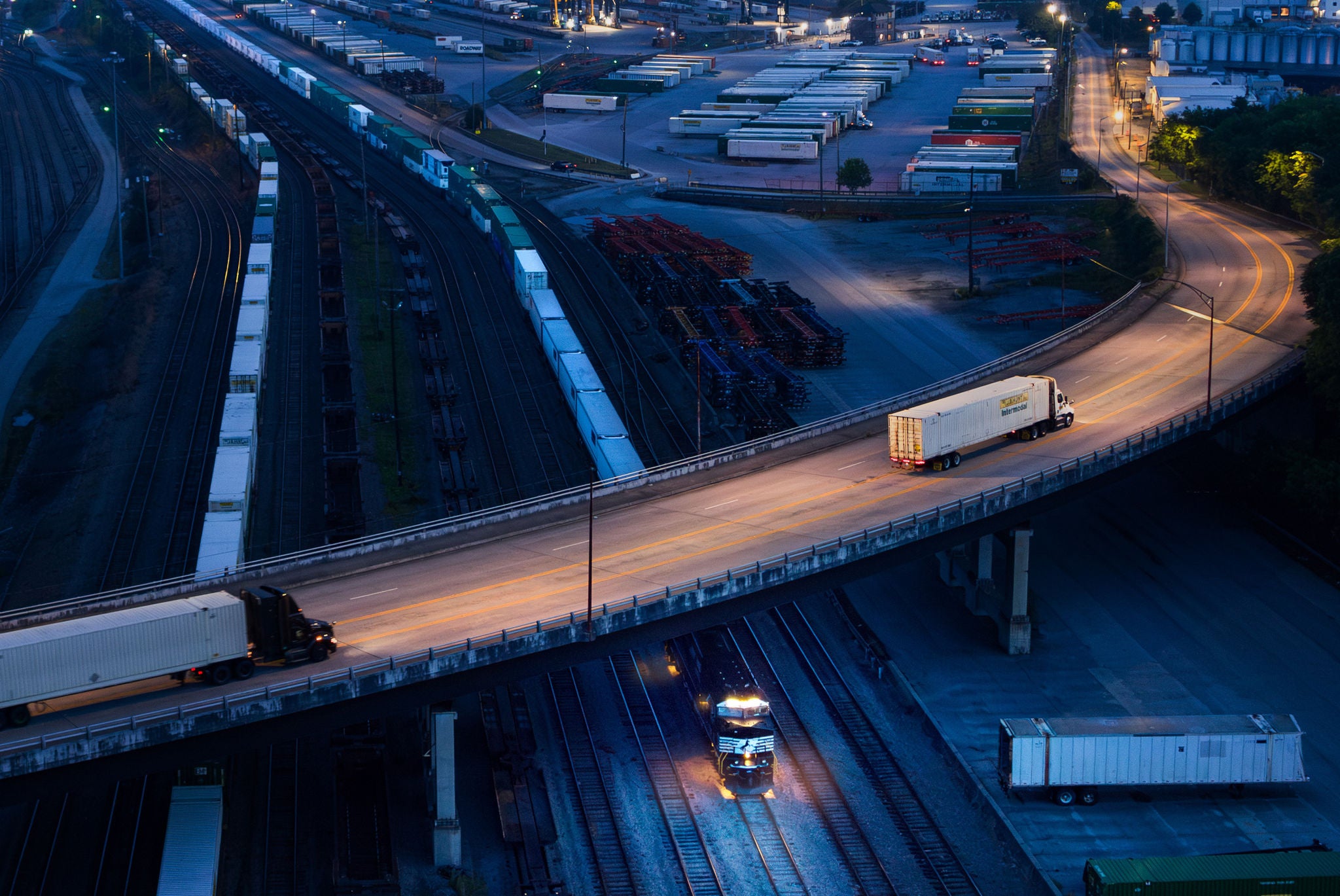 Aerial shot at night of rail freight industry featuring semi-trucks driving into train yard and two trains on rail tracks