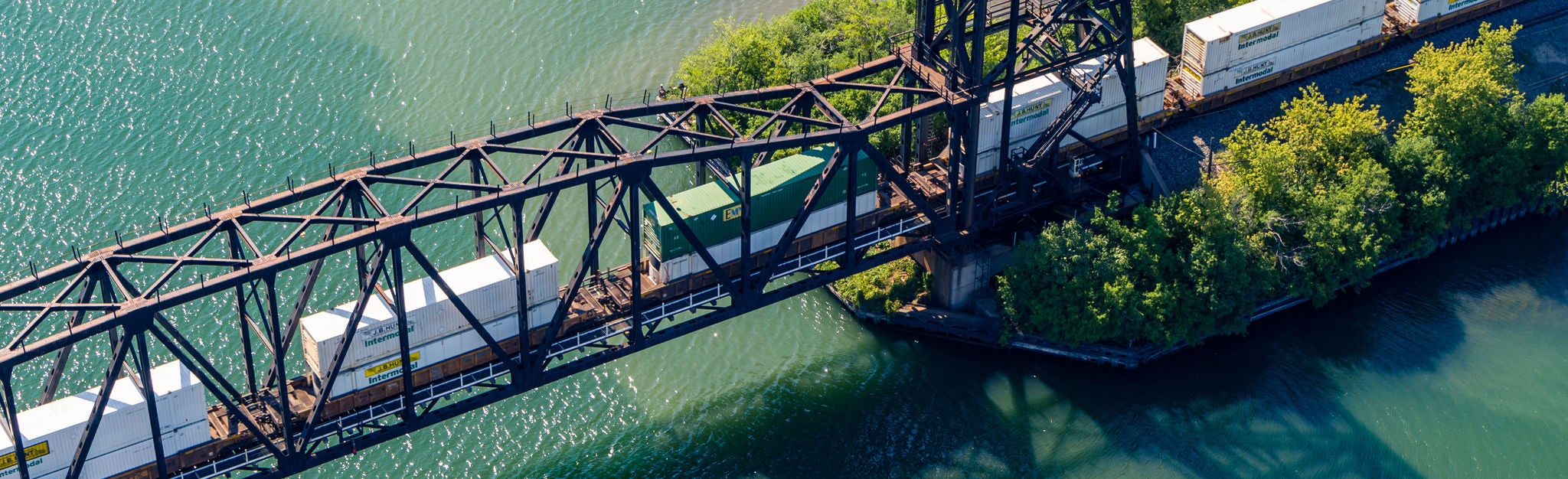 An aerial shot of an intermodal shipping train moving on a bridge over a river.